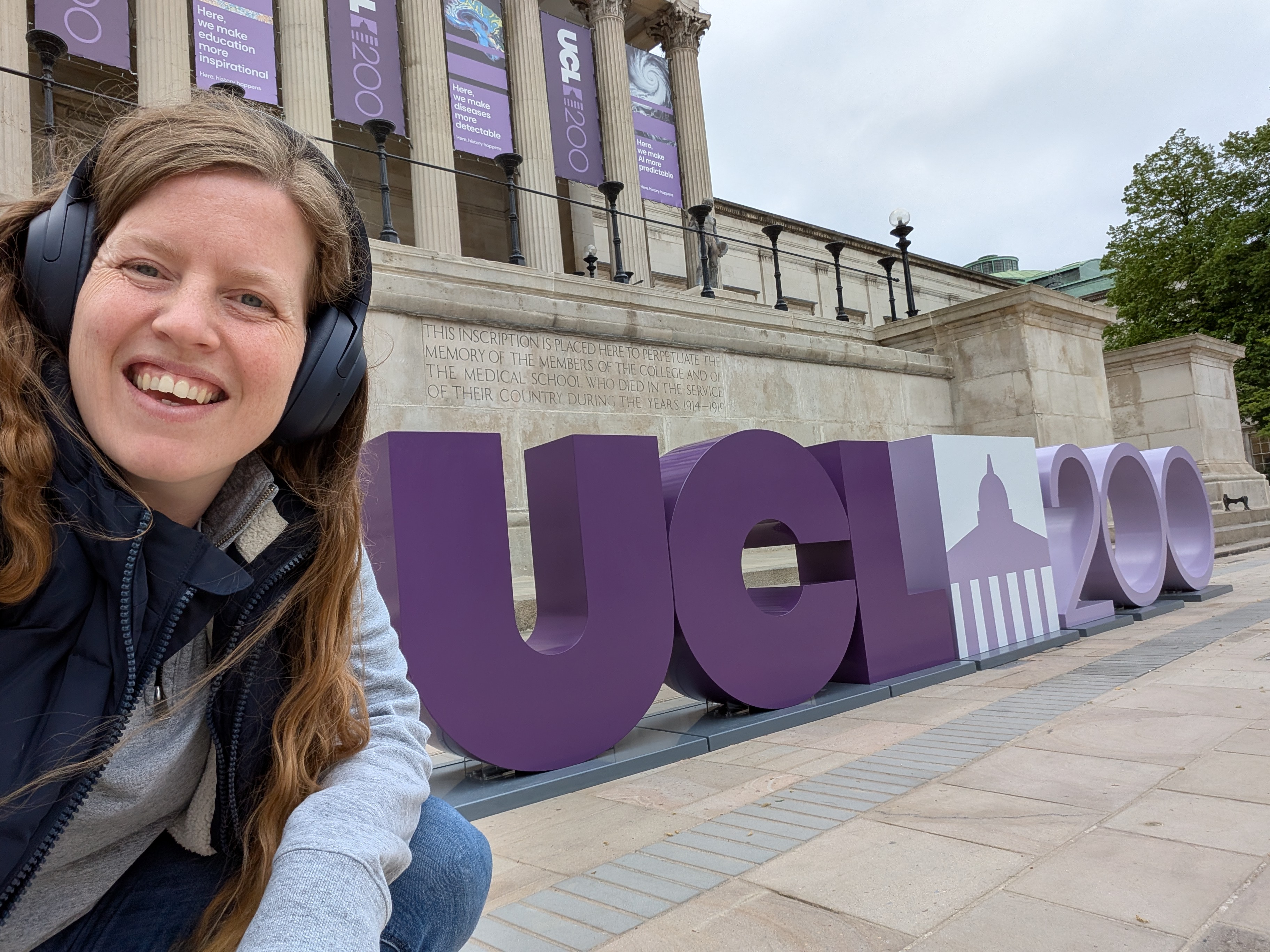 Research Podcasts' Director of Training and Production Krissie Brighty-Glover wearing headphones sat in front of a UCL 200 stand in UCL's quad on a recording day of the Bridge