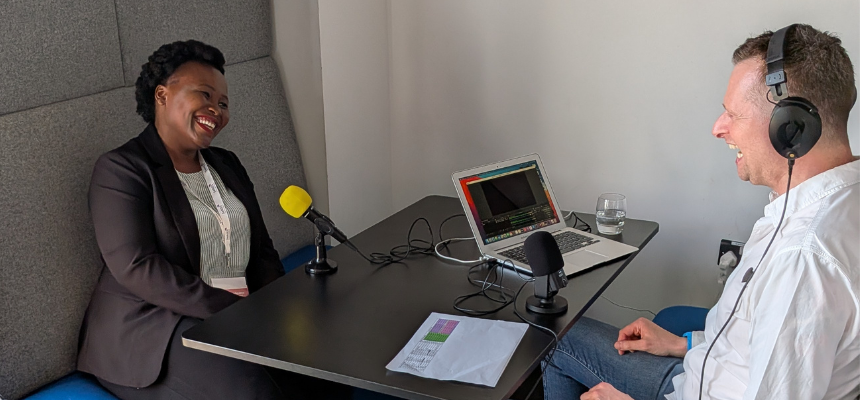 A high-angle photo capturing a Research Podcasts training session during the 2026 SeNSS conference at the University of Lincoln, where students recorded content for a podcast conference package. On the left, a Black female participant wearing a dark blazer, striped shirt, and conference lanyard sits on a high-backed grey booth bench, smiling broadly. Across the dark desk on the right, a trainer wearing a white button-down shirt and over-ear headphones laughs while facing her. On the table between them sits an open silver laptop displaying audio recording software, a glass of water, reference notes, and two tabletop microphones—one with a bright yellow foam windscreen positioned toward the speaker, and a black USB microphone near the laptop.